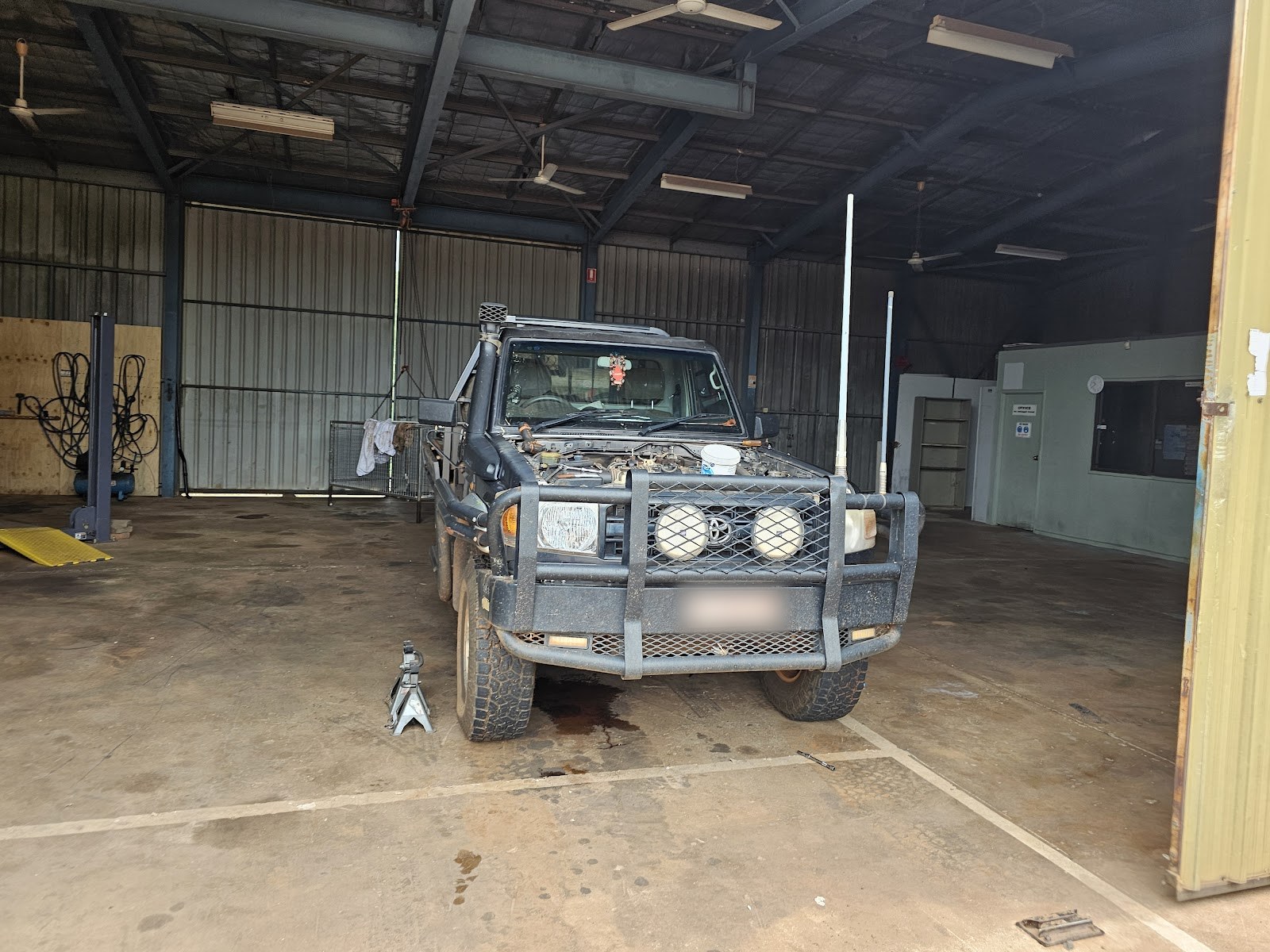 A black LandCruiser 70 ute with bullbar inside the TJ Auto Tech workshop, hoist and office behind